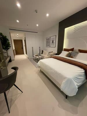 חדר מלון מודרני במלון Prime Residency 3, הכולל מיטה זוגית מסודרת, שמיכת פוך חומה, קיר דקורטיבי, שולחן וכסא, ספה, תמונות ממוסגרות, מנורות תקרה ומסדרון המוביל לדלת הכניסה. חדר מלון מודרני במלון Prime Residency 3, הכולל מיטה זוגית מסודרת, שמיכת פוך חומה, קיר דקורטיבי, שולחן וכסא, ספה, תמונות ממוסגרות, מנורות תקרה ומסדרון המוביל לדלת הכניסה.