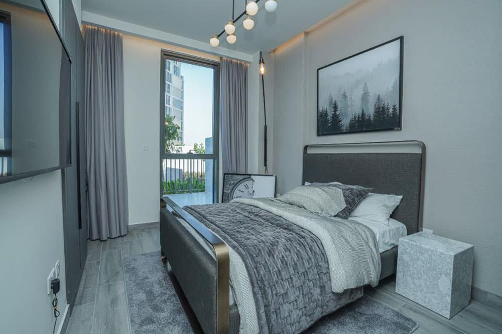 חדר שינה מודרני בפרויקט “Noor Midtown” של חברת Deyaar, המעוצב בגווני אפור, עם מיטה גדולה ומצעים בעלי מרקם, יצירת אמנות מופשטת המתארת יער מעל ראש המיטה, וילונות מהרצפה עד התקרה, דלת למרפסת וגופי תאורה עכשוויים, במרכז העיר דובאי. חדר שינה מודרני בפרויקט "Noor Midtown" של חברת Deyaar, המעוצב בגווני אפור, עם מיטה גדולה ומצעים בעלי מרקם, יצירת אמנות מופשטת המתארת יער מעל ראש המיטה, וילונות מהרצפה עד התקרה, דלת למרפסת וגופי תאורה עכשוויים, במרכז העיר דובאי.