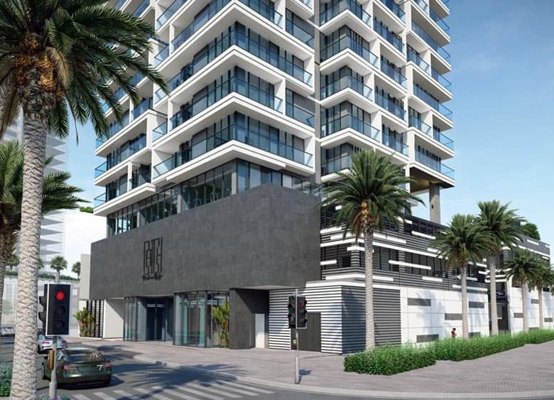 Жилой комплекс Catch Residences — это современное многоэтажное здание со стеклянными балконами, окруженное пальмами и зеленью, расположенное на перекрестке Реховот. На фотографии — автомобиль, ожидающий на красный свет под ясным голубым небом.