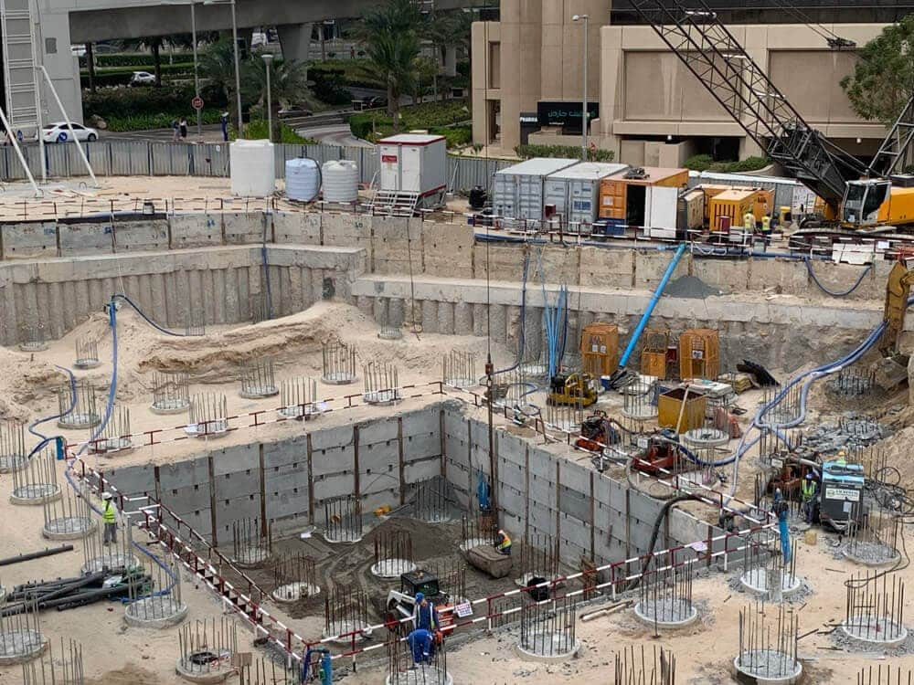אתר בנייה עם בור חפירה גדול, עמודי מוטות מברזל ותבניות בטון. במקום נמצאים פועלים לבושים בציוד בטיחות, ובסביבתו נראים כלים מכניים, כבלים ומבנים זמניים. ברקע נראים מבנים עירוניים. אתר בנייה עם בור חפירה גדול, עמודי מוטות מברזל ותבניות בטון. במקום נמצאים פועלים לבושים בציוד בטיחות, ובסביבתו נראים כלים מכניים, כבלים ומבנים זמניים. ברקע נראים מבנים עירוניים.