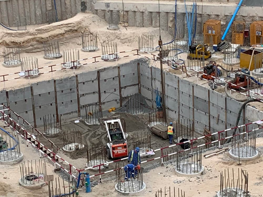 אתר בנייה שבו חפירה מלבנית עמוקה המוקפת בקירות בטון ועמודי מוטות פלדה; בתוך הבור נמצאים דחפור קטן ושני פועלים לבושים באפודי בטיחות, ובסביבתו פזורים כלי בנייה. אתר בנייה שבו חפירה מלבנית עמוקה המוקפת בקירות בטון ועמודי מוטות פלדה; בתוך הבור נמצאים דחפור קטן ושני פועלים לבושים באפודי בטיחות, ובסביבתו פזורים כלי בנייה.