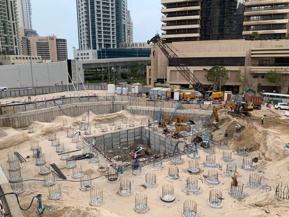 אתר בנייה שבו נראים פועלים, מכונות ועמודי מוטות מברזל חשופים בשטח חפירה נרחב, המוקף בבניינים גבוהים ובציוד, המעיד על השלבים הראשונים של עבודות הנחת היסודות. אתר בנייה שבו נראים פועלים, מכונות ועמודי מוטות מברזל חשופים בשטח חפירה נרחב, המוקף בבניינים גבוהים ובציוד, המעיד על השלבים הראשונים של עבודות הנחת היסודות.