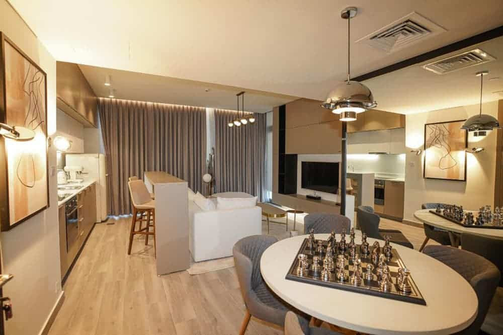 סלון ופינת אוכל מודרניים בפרויקט “Noor Midtown” של חברת Deyaar, הכוללים רצפות עץ בהירות, ספה לבנה, פינת מטבח, יצירות אמנות על הקירות, שולחנות עגולים עם ערכות שחמט וגופי תאורה עכשוויים. וילונות מהרצפה עד התקרה מכסים את החלונות הגדולים ברקע. סלון ופינת אוכל מודרניים בפרויקט "Noor Midtown" של חברת Deyaar, הכוללים רצפות עץ בהירות, ספה לבנה, פינת מטבח, יצירות אמנות על הקירות, שולחנות עגולים עם ערכות שחמט וגופי תאורה עכשוויים. וילונות מהרצפה עד התקרה מכסים את החלונות הגדולים ברקע.