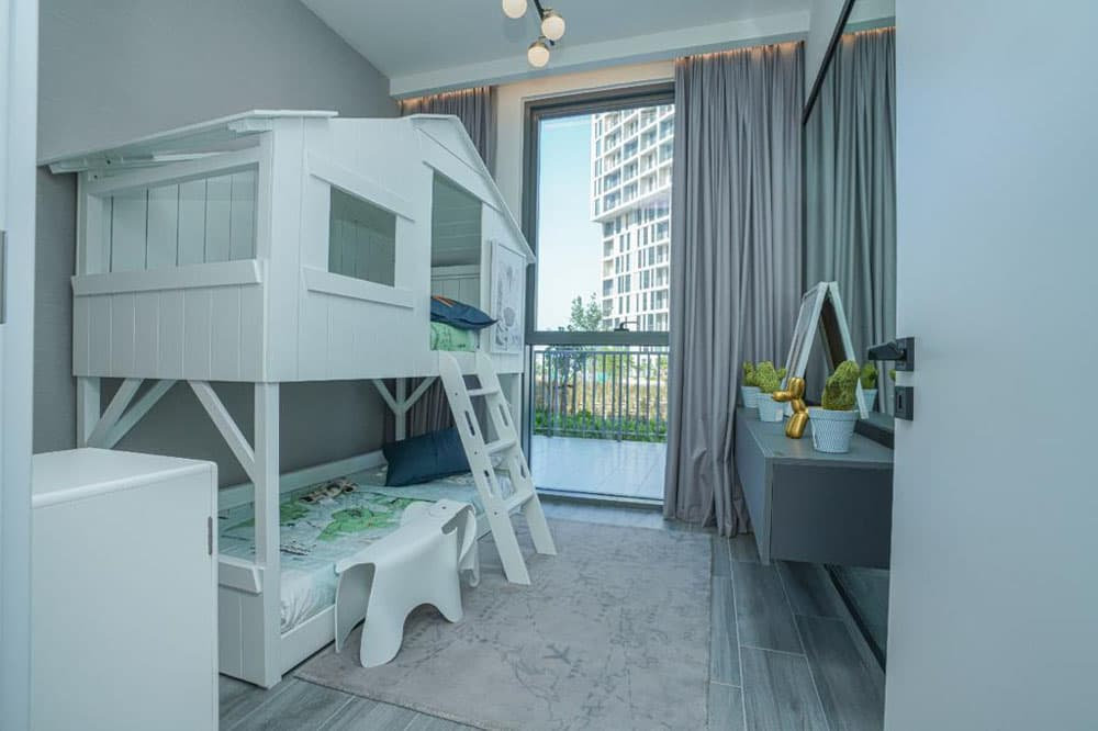 חדר שינה מודרני לילדים בפרויקט “Noor Midtown” של חברת Deyaar, הכולל מיטת קומתיים לבנה, שולחן כתיבה קטן עם מראה וקישוטים, חלון גדול עם וילונות אפורים, ונוף של בניינים גבוהים וצמחייה. גווני תכלת ואפור משלימים את האווירה הרעננה של החדר. חדר שינה מודרני לילדים בפרויקט "Noor Midtown" של חברת Deyaar, הכולל מיטת קומתיים לבנה, שולחן כתיבה קטן עם מראה וקישוטים, חלון גדול עם וילונות אפורים, ונוף של בניינים גבוהים וצמחייה. גווני תכלת ואפור משלימים את האווירה הרעננה של החדר.