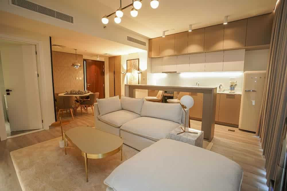 סלון ומטבח מודרניים בתכנון פתוח בפרויקט “Noor Midtown” של חברת Deyaar, הכוללים ספות בצבע בז’, שולחן קפה עם נגיעות זהב, תאורה תקרתית, ארונות מובנים ופינת אוכל נעימה. החלל משדר חמימות ומראה עכשווי. סלון ומטבח מודרניים בתכנון פתוח בפרויקט "Noor Midtown" של חברת Deyaar, הכוללים ספות בצבע בז', שולחן קפה עם נגיעות זהב, תאורה תקרתית, ארונות מובנים ופינת אוכל נעימה. החלל משדר חמימות ומראה עכשווי.