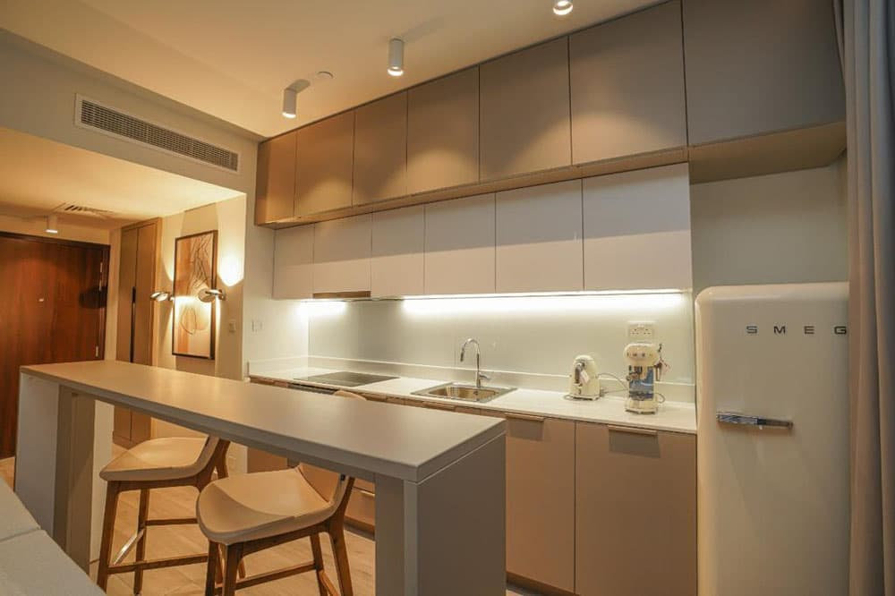 מטבח מודרני בפרויקט “Noor Midtown” של חברת Deyaar, הכולל ארונות בצבע בז’, אי ארוך עם שני כסאות בר מעץ, תאורה תחתונה לארונות, מקרר SMEG לבן ומיקסר ליד הכיור. התאורה החמימה והעיצוב המינימליסטי יוצרים אווירה נעימה בסגנון מידטאון דובאי. מטבח מודרני בפרויקט "Noor Midtown" של חברת Deyaar, הכולל ארונות בצבע בז', אי ארוך עם שני כסאות בר מעץ, תאורה תחתונה לארונות, מקרר SMEG לבן ומיקסר ליד הכיור. התאורה החמימה והעיצוב המינימליסטי יוצרים אווירה נעימה בסגנון מידטאון דובאי.