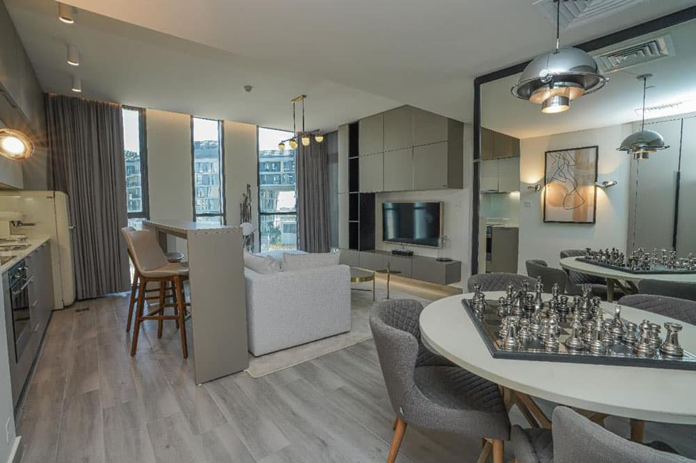 סלון ומטבח מודרניים בתכנון פתוח בפרויקט “Deyaar Noor Midtown”, המעוצבים בגוונים אפורים וניטרליים, עם ספה בהירה, טלוויזיה עם מסך שטוח, שולחן אוכל עם משטח דמוי לוח שחמט, כסאות בר, חלונות גדולים עם וילונות ותאורה עכשווית. סלון ומטבח מודרניים בתכנון פתוח בפרויקט "Deyaar Noor Midtown", המעוצבים בגוונים אפורים וניטרליים, עם ספה בהירה, טלוויזיה עם מסך שטוח, שולחן אוכל עם משטח דמוי לוח שחמט, כסאות בר, חלונות גדולים עם וילונות ותאורה עכשווית.